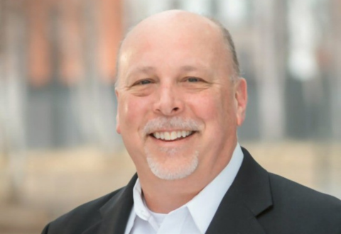 The image is of a bald white man with a grey goatee smiling against a blurred background of a city. He is wearing a white button-up under a black suit jacket.